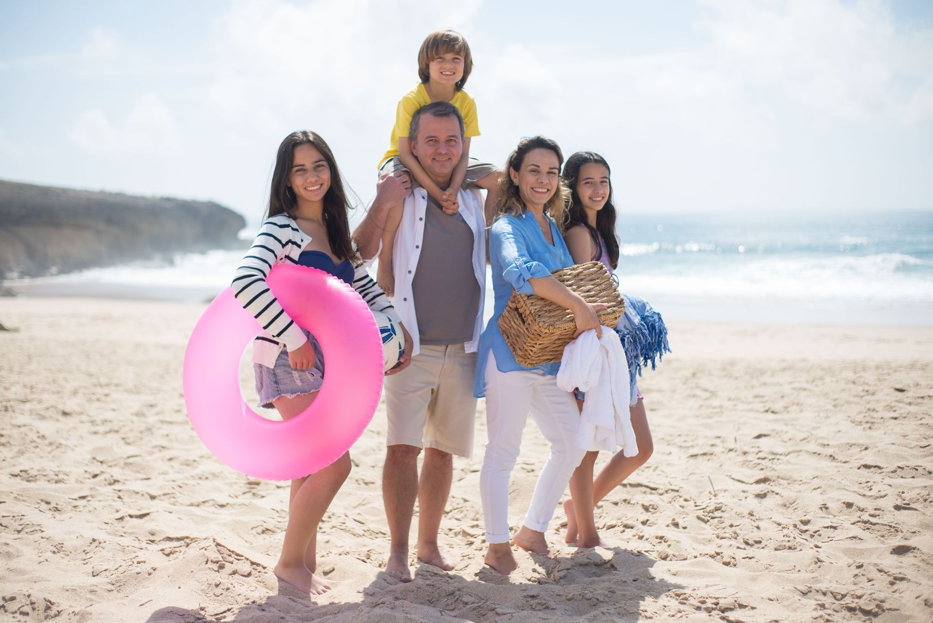 Group of people on a sunny beach, one holding a pink swim ring and another a child on her shoulders.