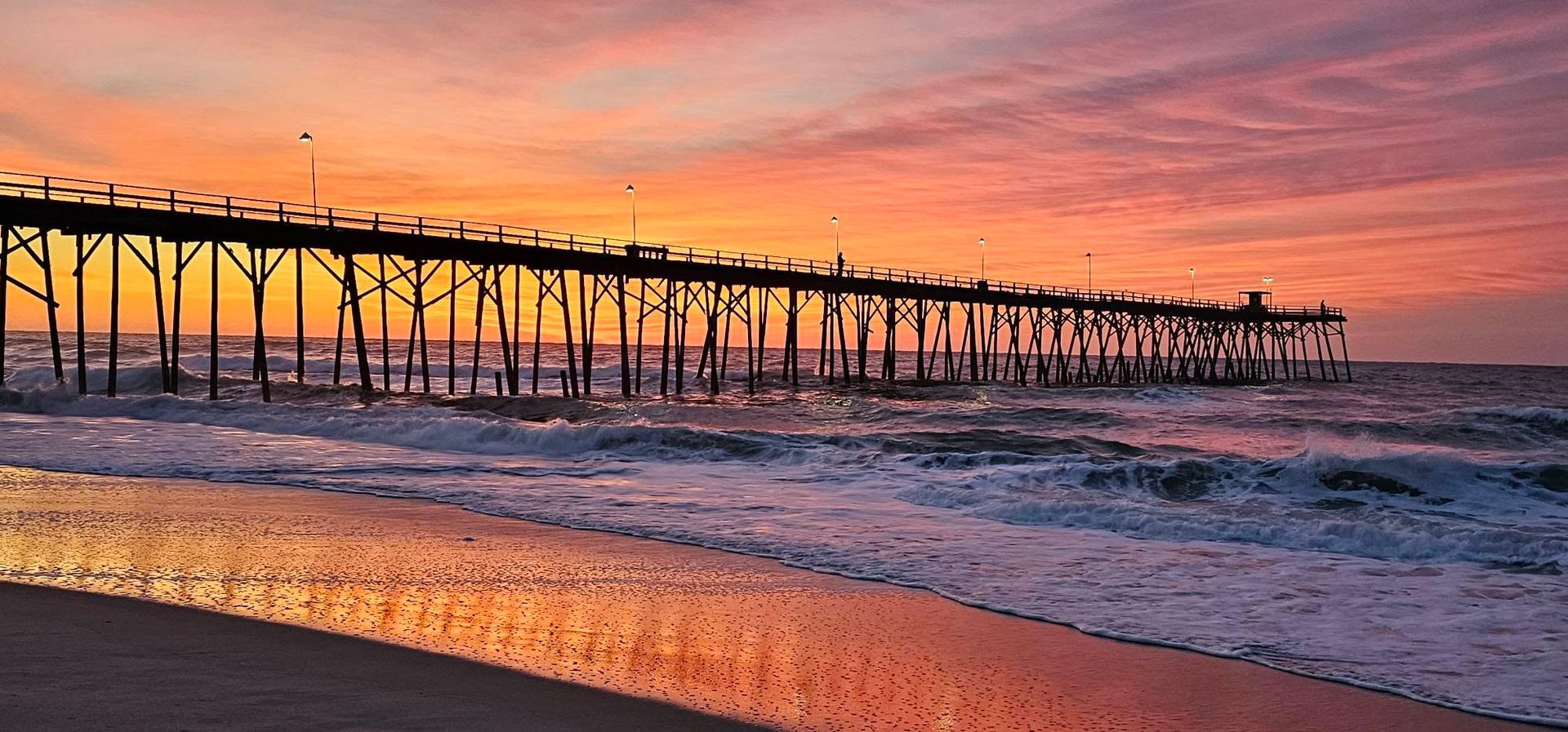Sunset over a wooden pier above ocean waves, with orange and pink sky reflecting on the water