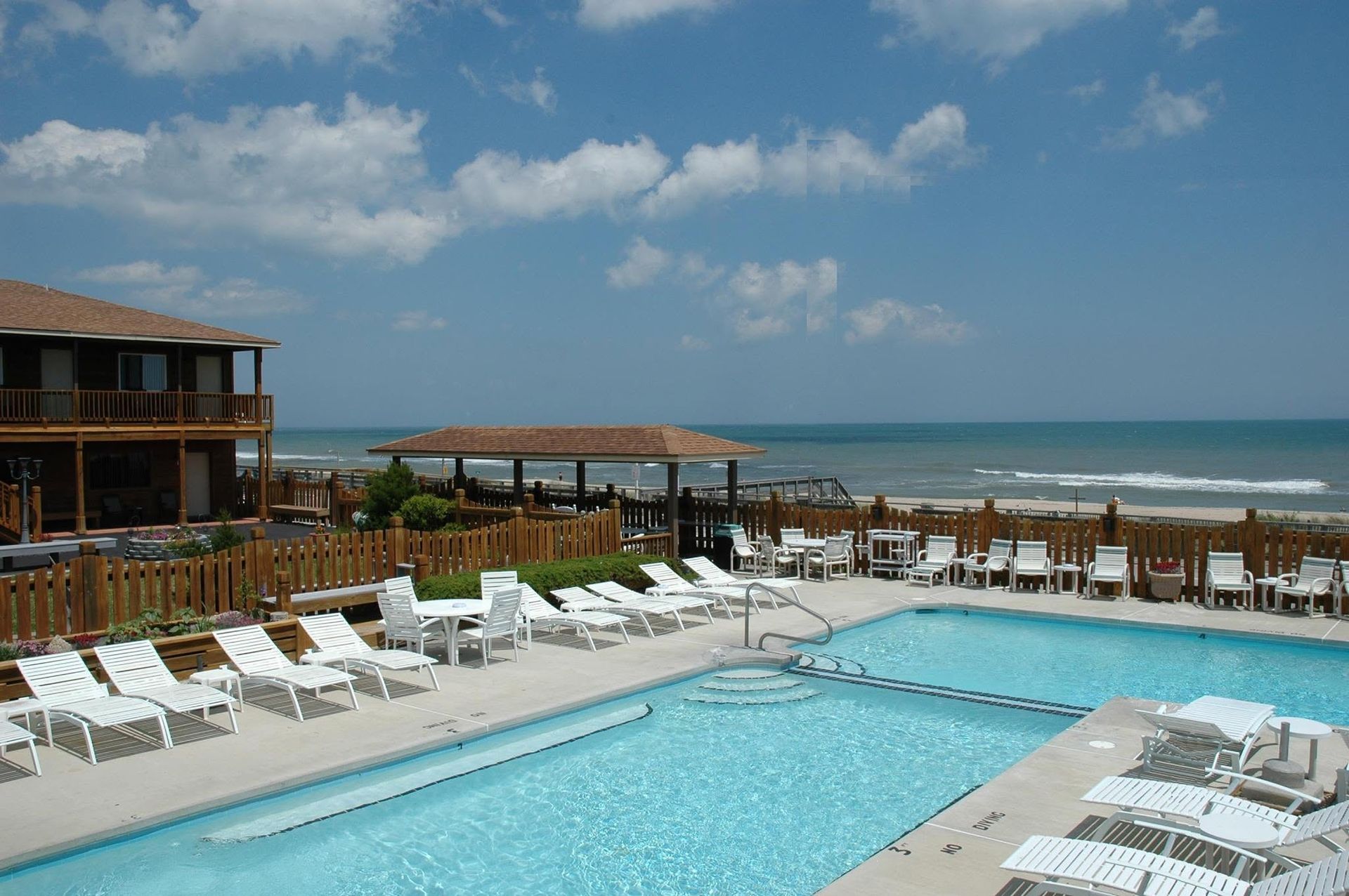 Resort pool with white lounge chairs, ocean view, and blue sky with clouds