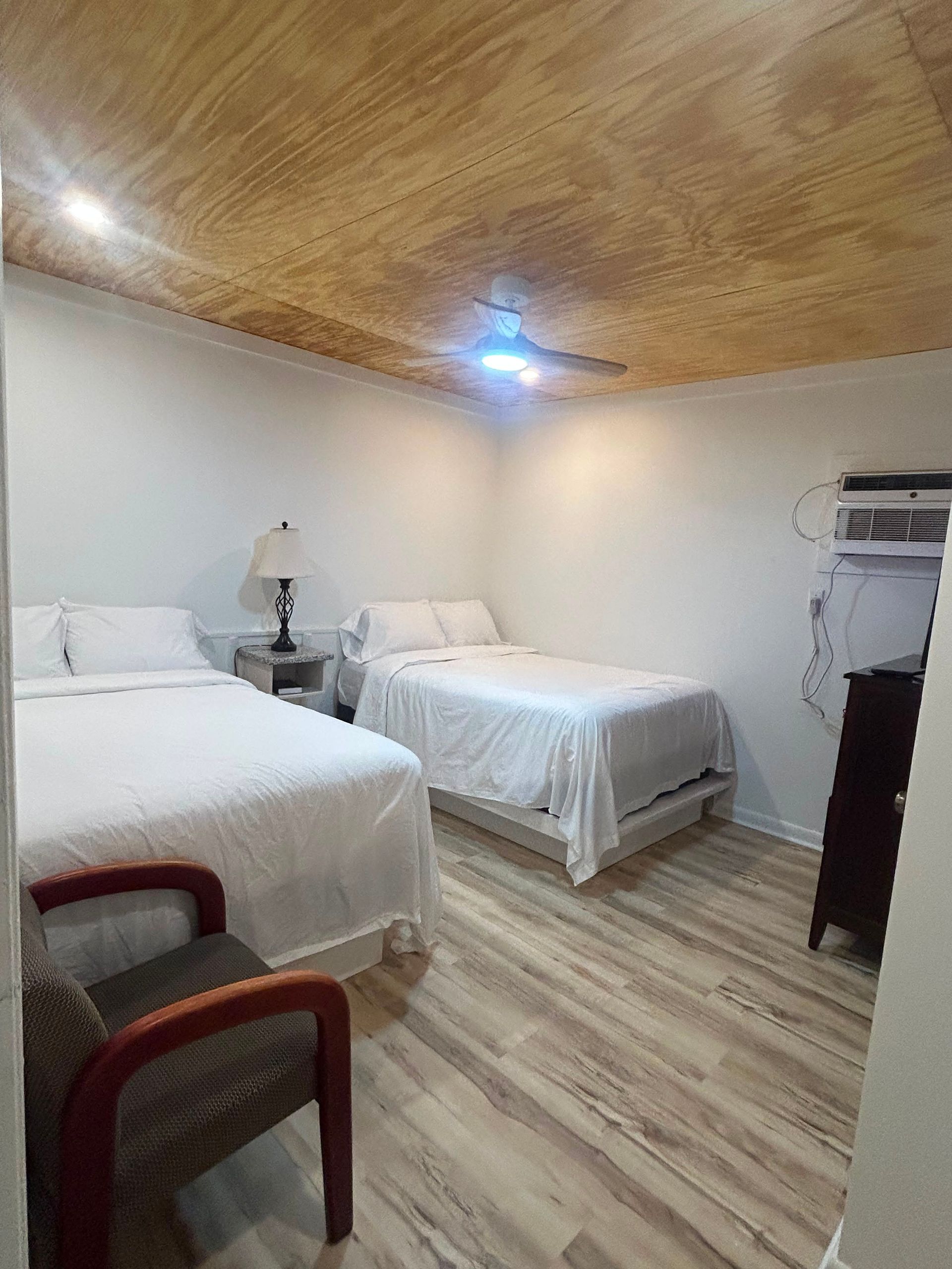 Small bedroom with two twin beds, white walls, wood ceiling, and a chair in the foreground.
