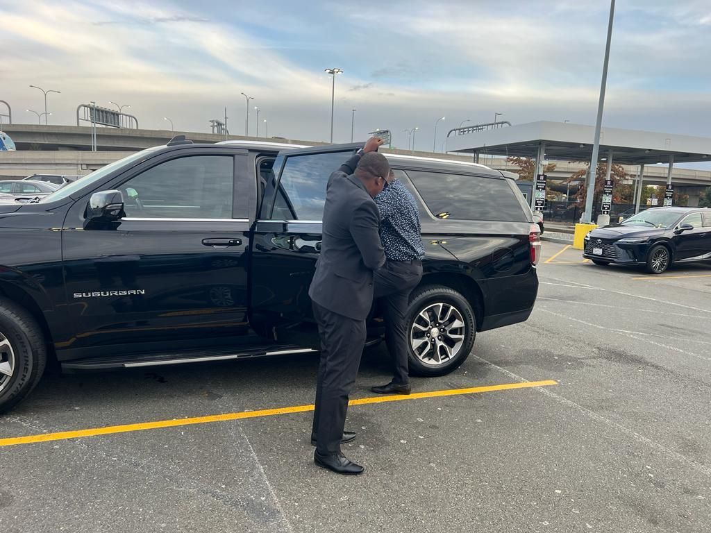 Two people in suits stand by a black SUV with an open door in a paved parking lot near a gas station.
