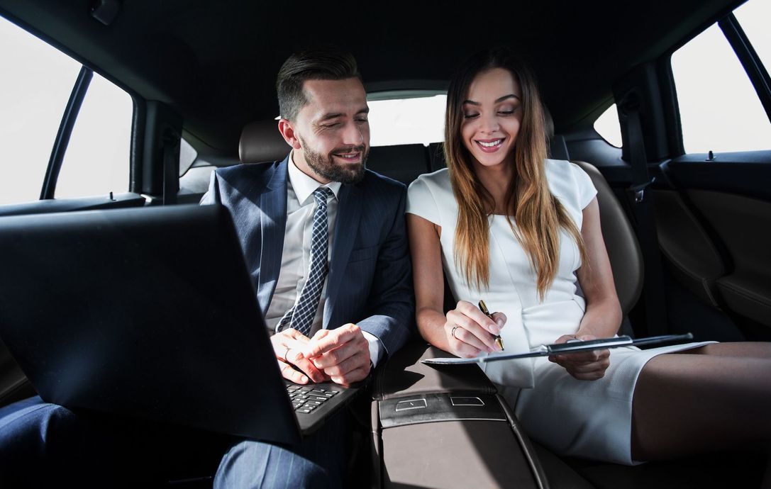 A man and a woman in professional attire sitting in the back of a car, reviewing documents and working on a laptop.