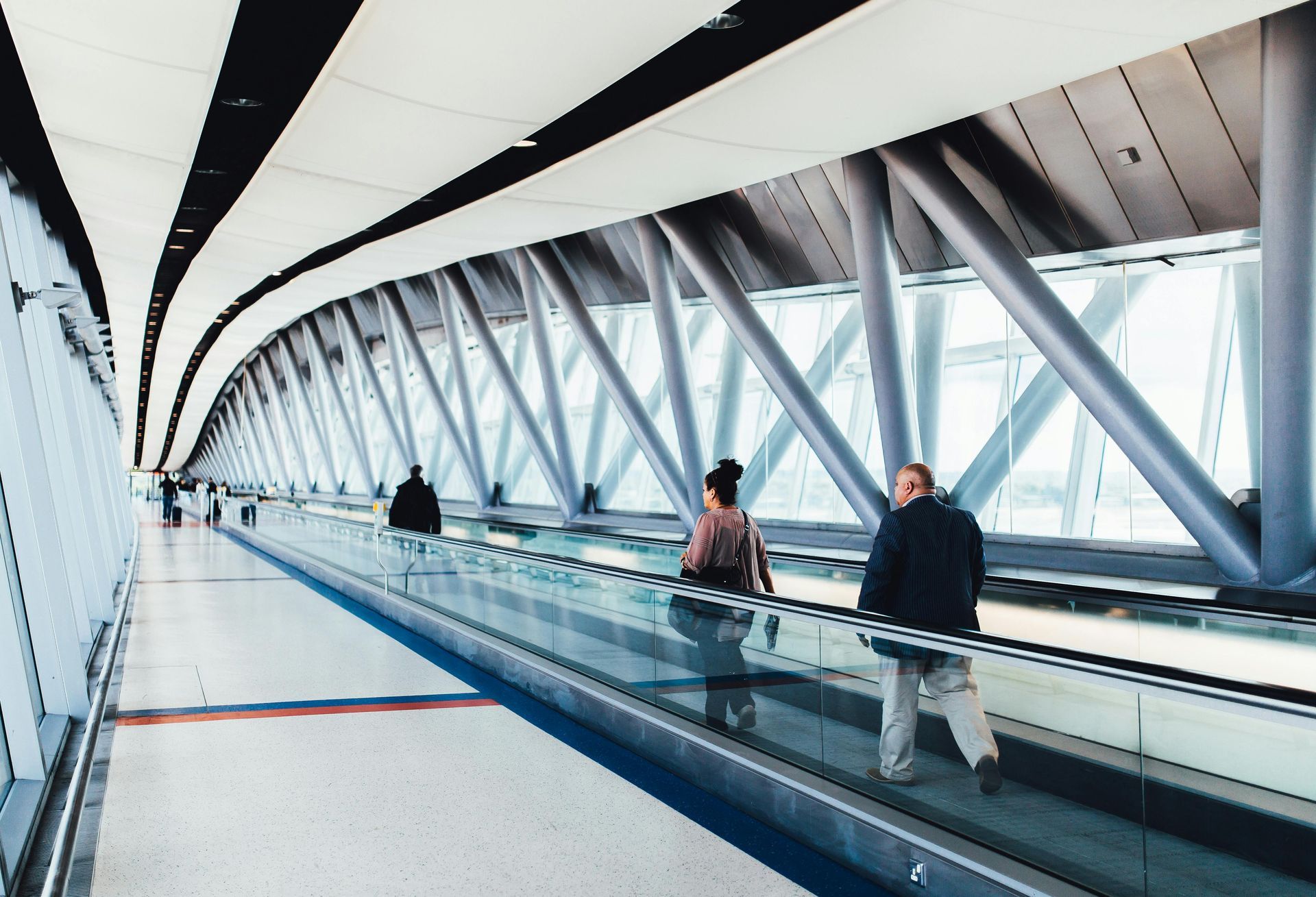 People walking on a moving walkway inside a modern, glass-walled airport terminal with a curved, industrial ceiling.
