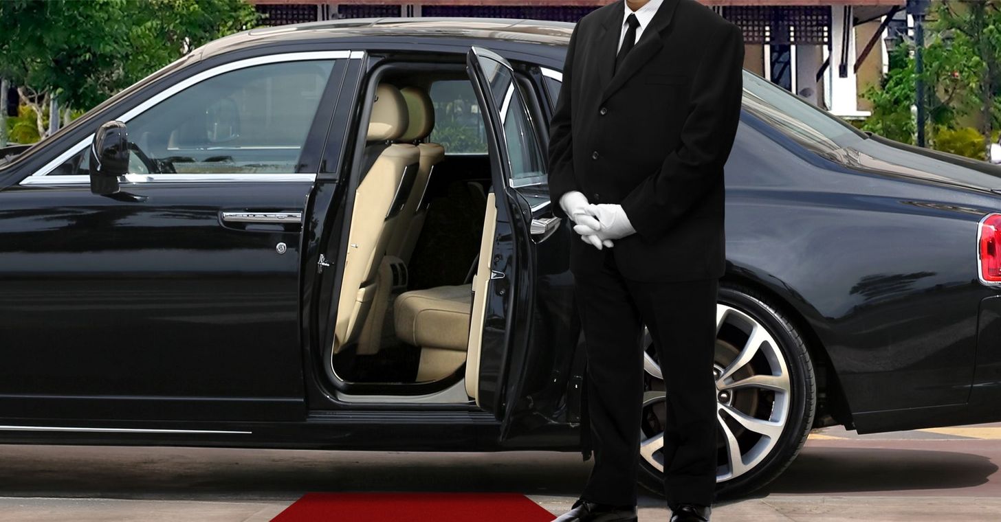 A uniformed chauffeur in white gloves stands next to a black luxury car with an open door and red carpet.