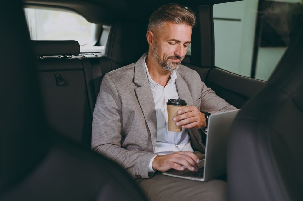A person in a business blazer works on a laptop while holding a coffee cup in the back seat of a car.