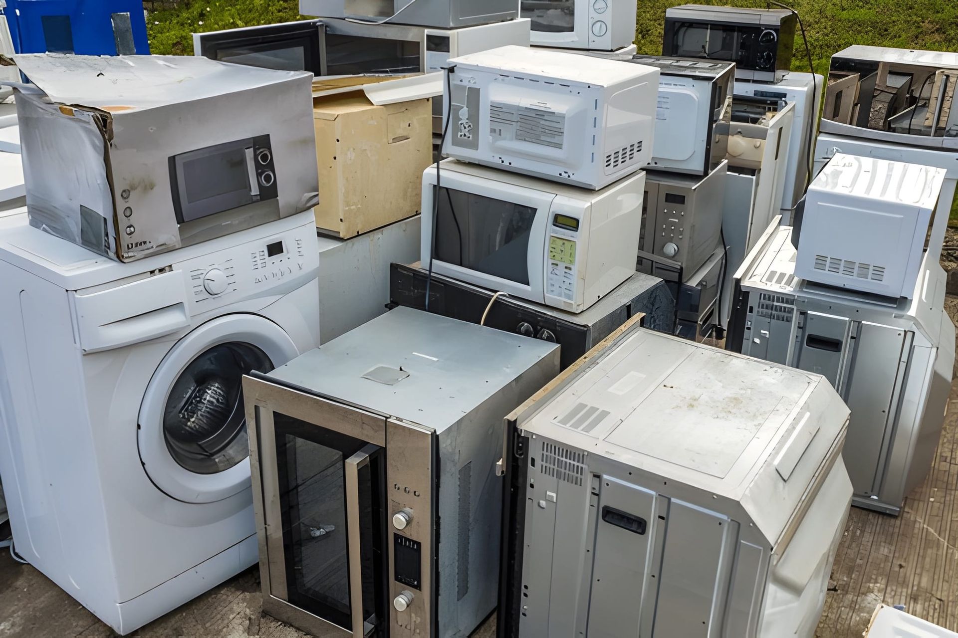 Pile of discarded appliances including a washing machine and microwaves, outside.