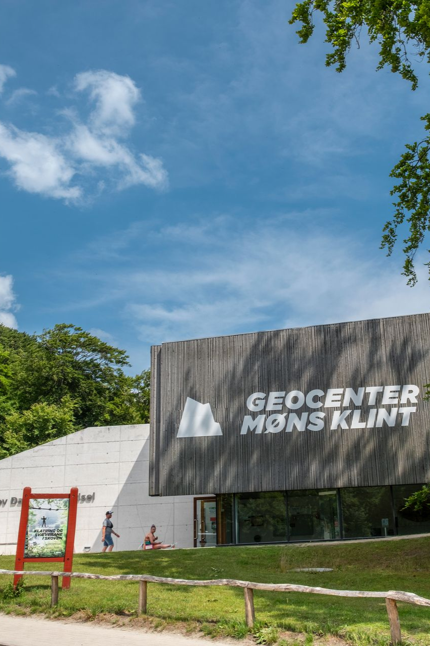 Geocenter Møns Klint bygning med en mørk træfacade under en blå himmel og grøn græsplæne