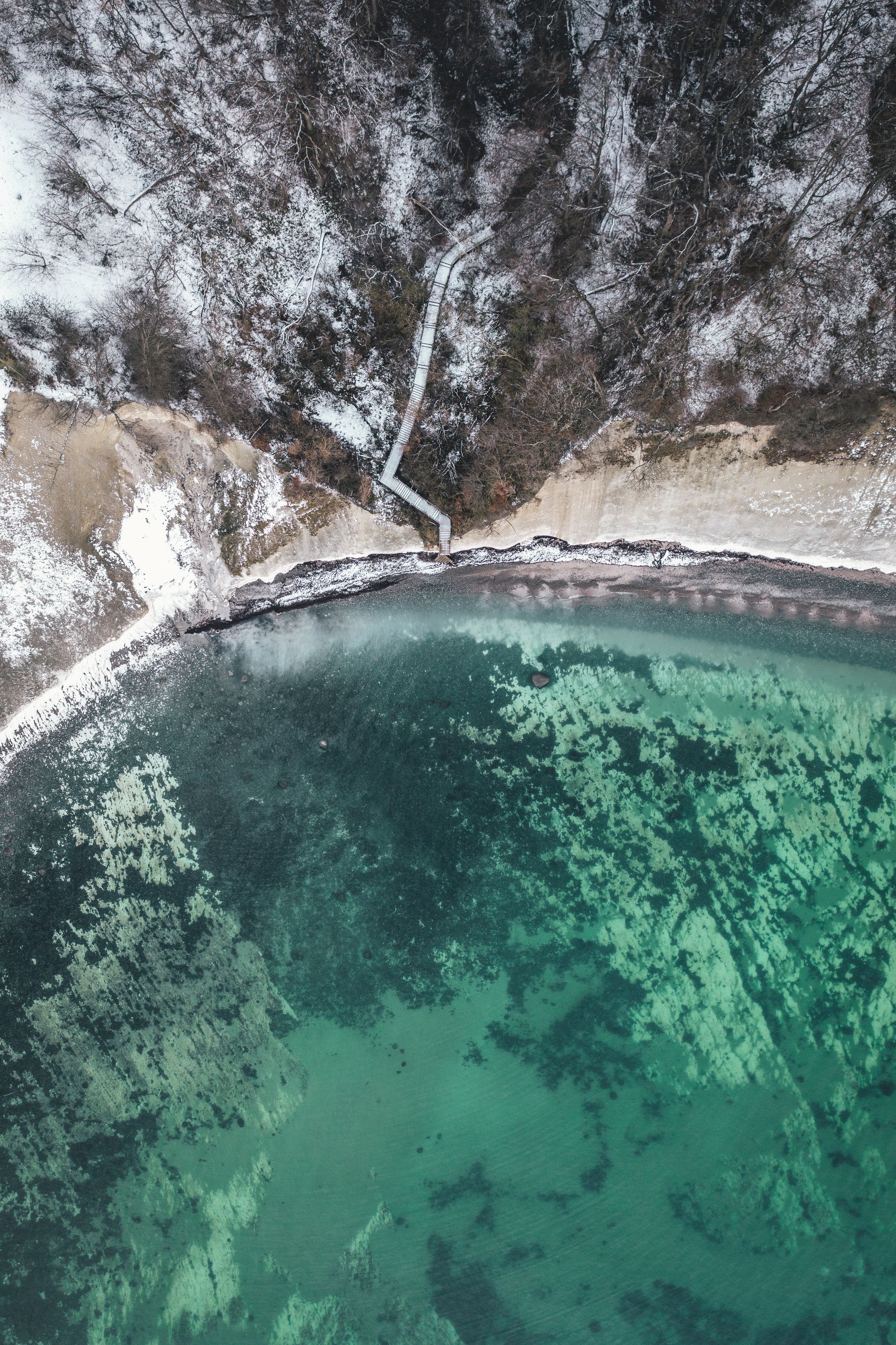 Luftfoto af en turkis kystlinje med mørke klipper og en smal hvid sandstrand