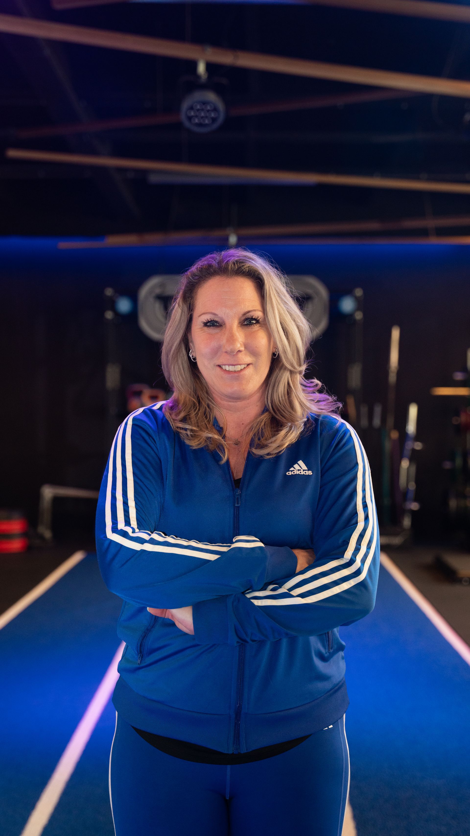 Vrouw in een blauw Adidas-jack met gekruiste armen in een sportschool.