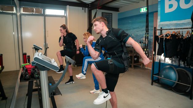 Mensen in EHBO-pakken oefenen binnen: stappen, knieën optrekken. Sportzaal met apparatuur, blauwe muur.