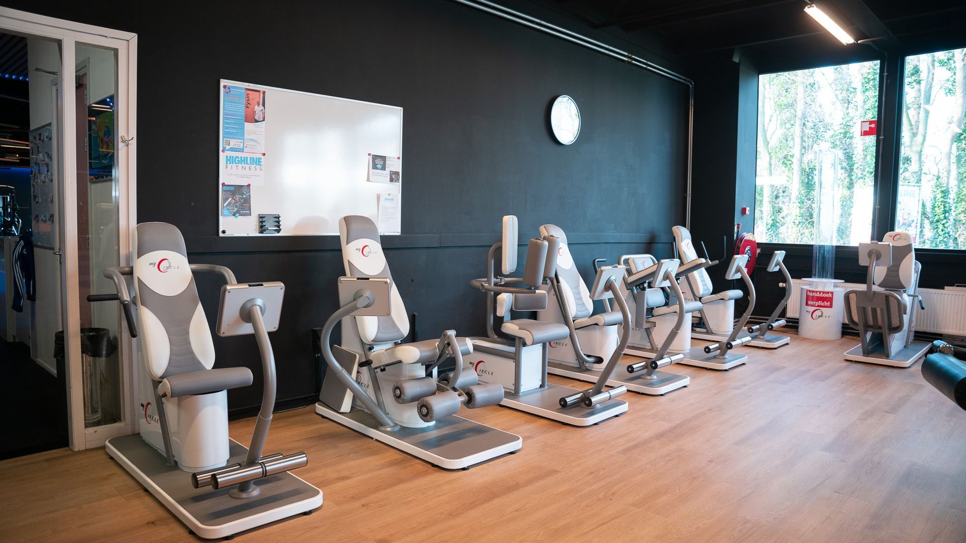 Interieur van een sportschool met fitnessapparaten op een rij, een zwarte muur, een whiteboard en een raam met groen.