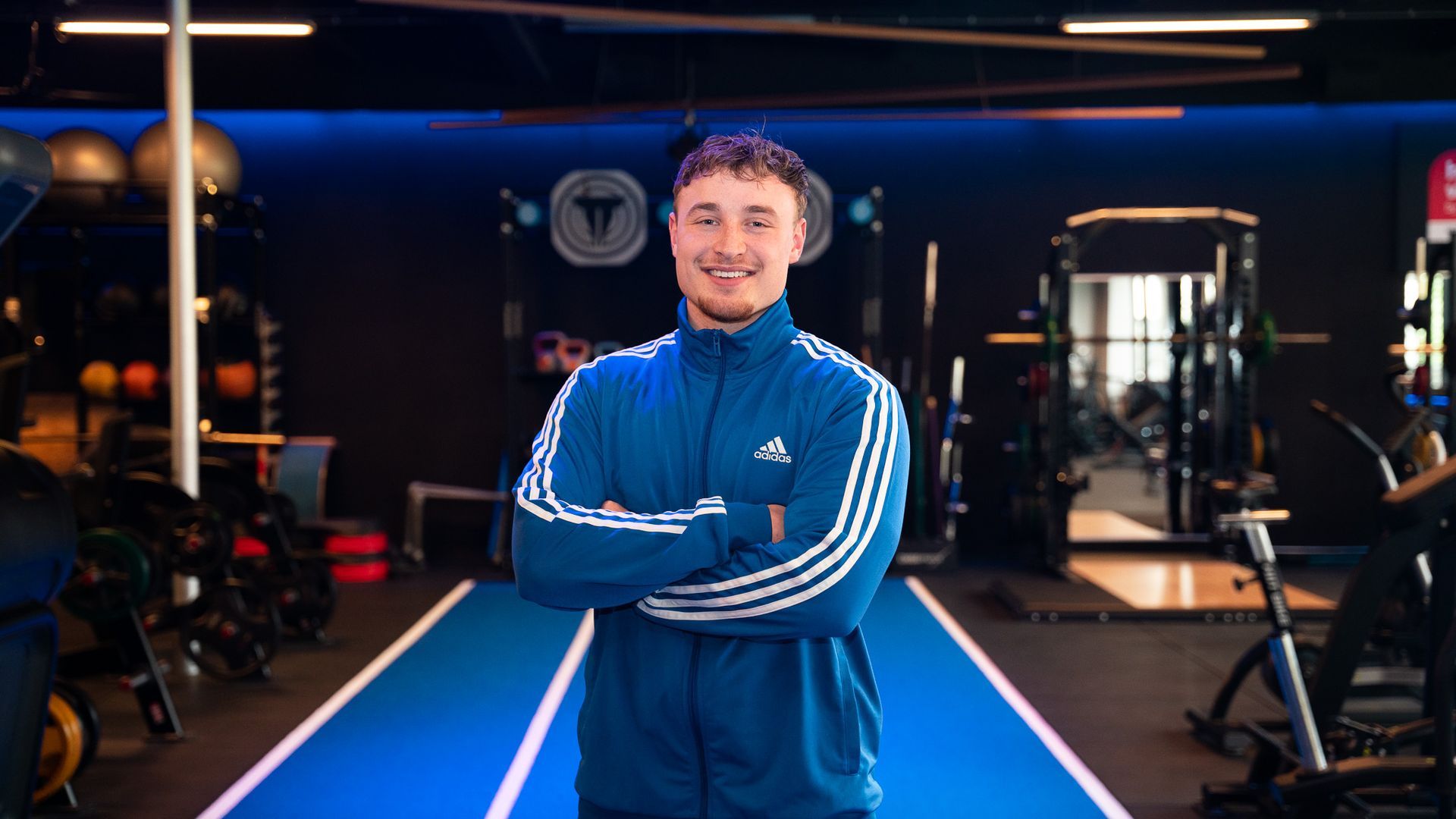 Man in een blauw Adidas-trainingspak met gekruiste armen, lachend in een sportschool.