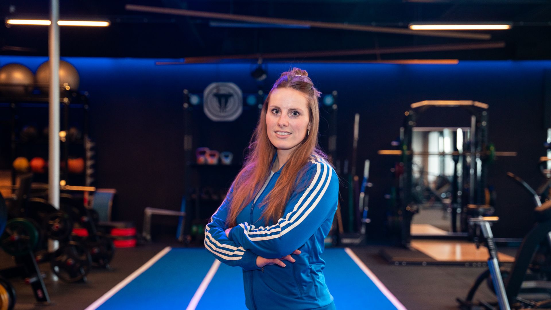 Een vrouw in een blauw jasje, met haar armen over elkaar, staat in een sportschool met fitnessapparatuur.