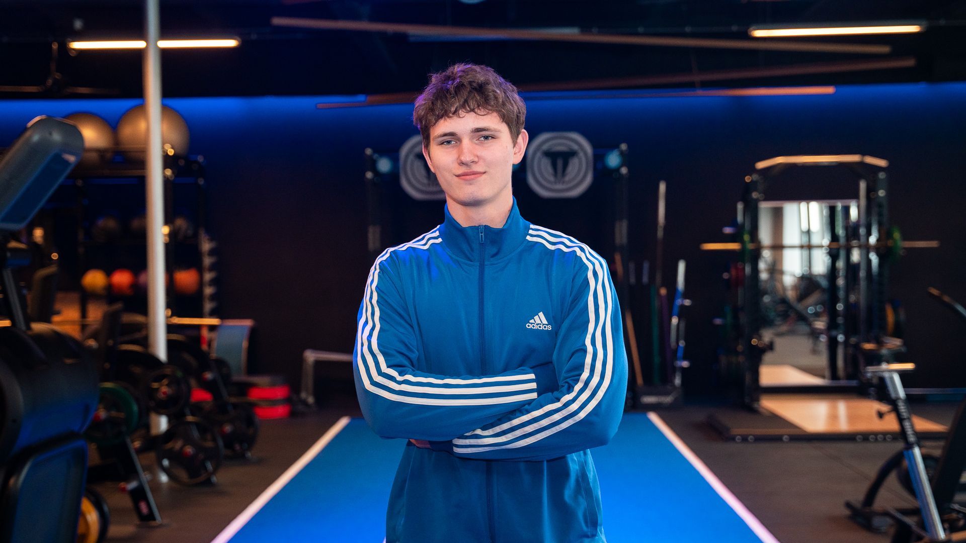 Jongeman in een blauw Adidas-trainingsjack, met gekruiste armen, staat in een sportschool.