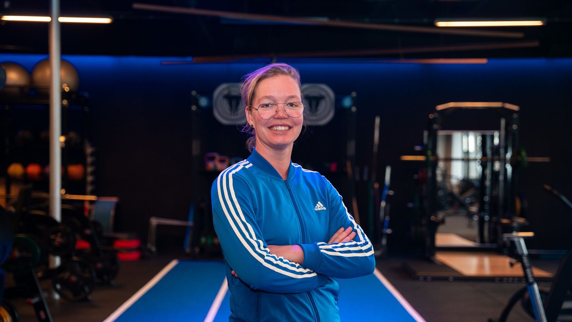 Een vrouw in een blauw trainingspak lacht in de sportschool, met haar armen over elkaar.
