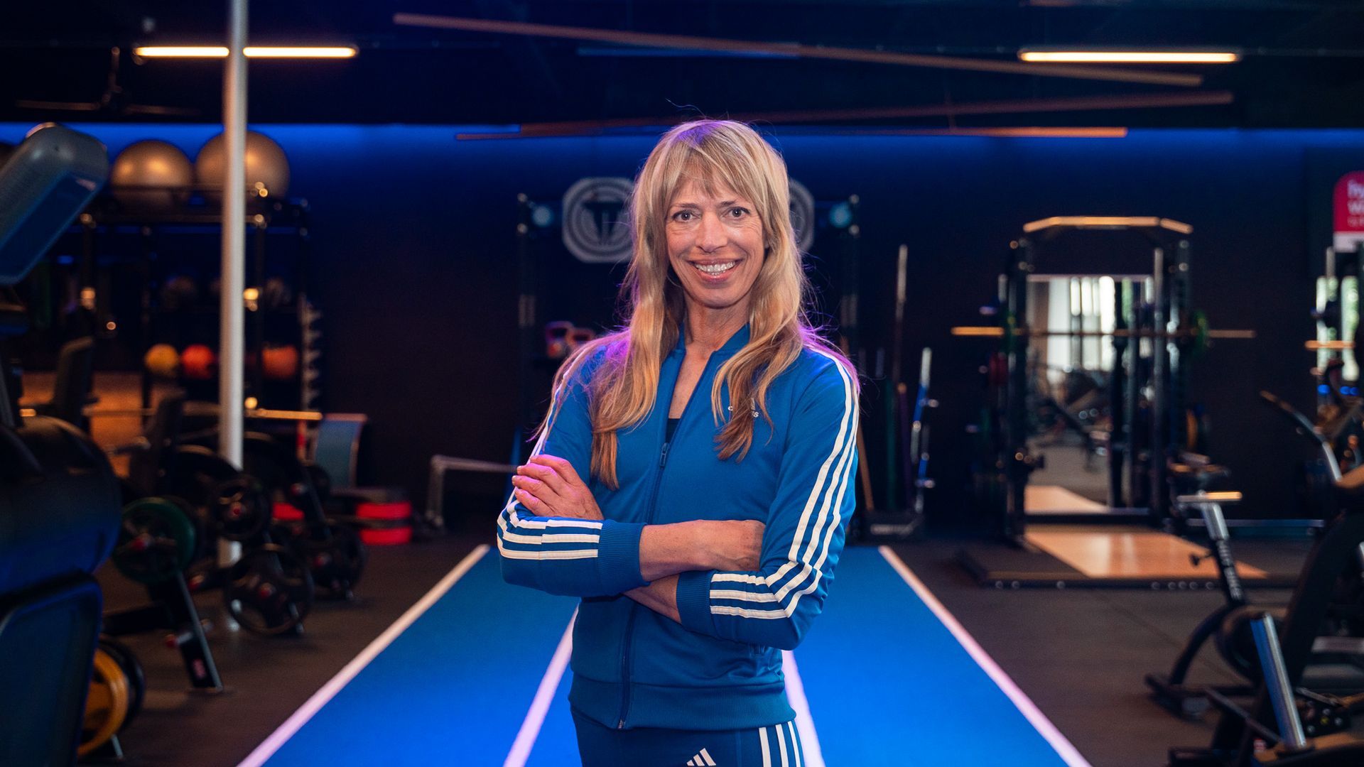 Vrouw in een blauw Adidas-jack met gekruiste armen in een sportschool.