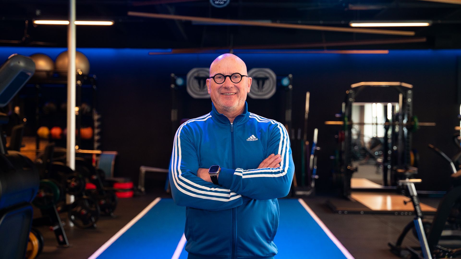 Kale man in een blauw Adidas-trainingspak, armen over elkaar, lachend in een sportschool.