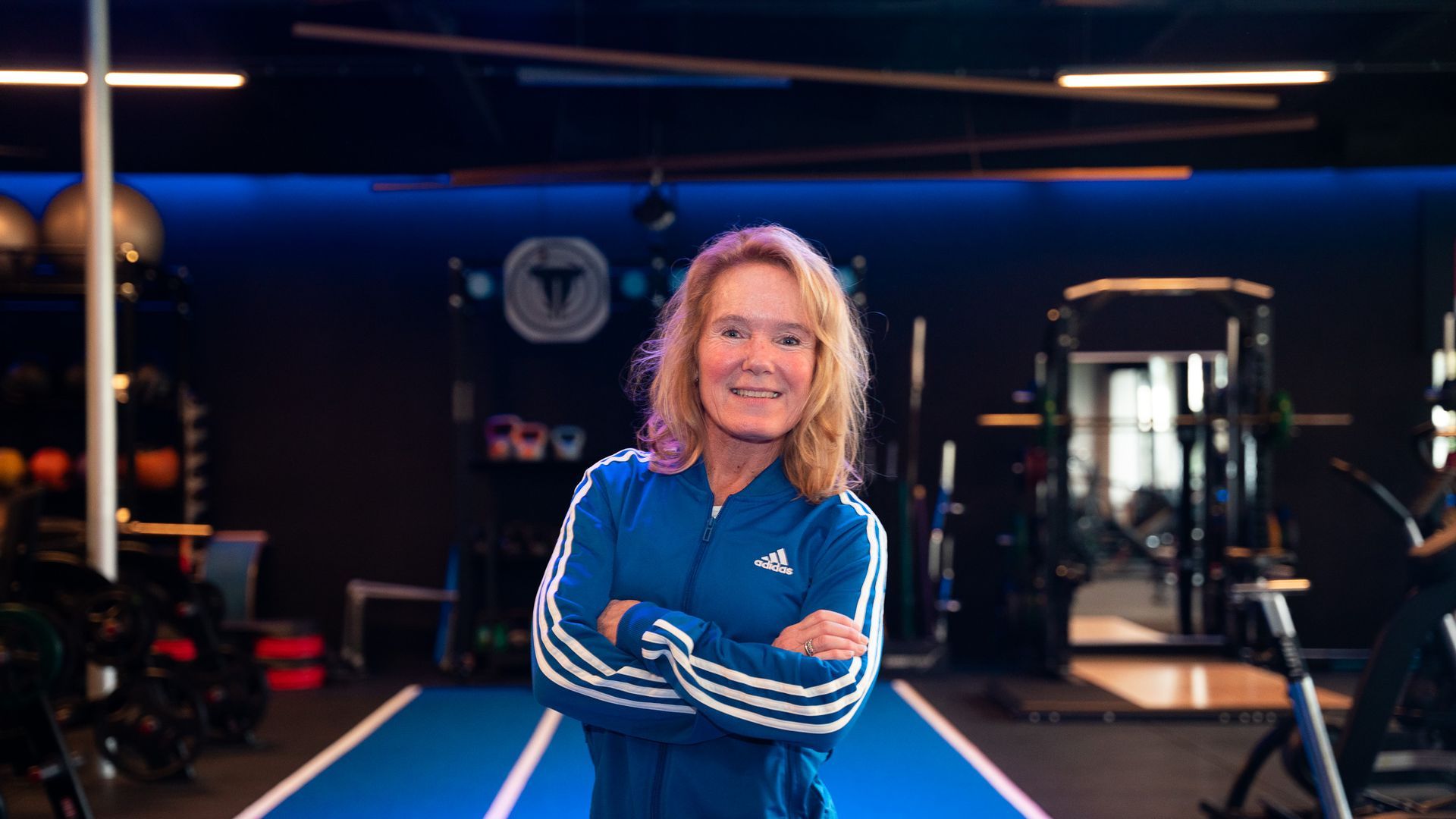 Een vrouw in een blauw trainingsjack, met haar armen over elkaar, staat in een sportschool met fitnessapparatuur.