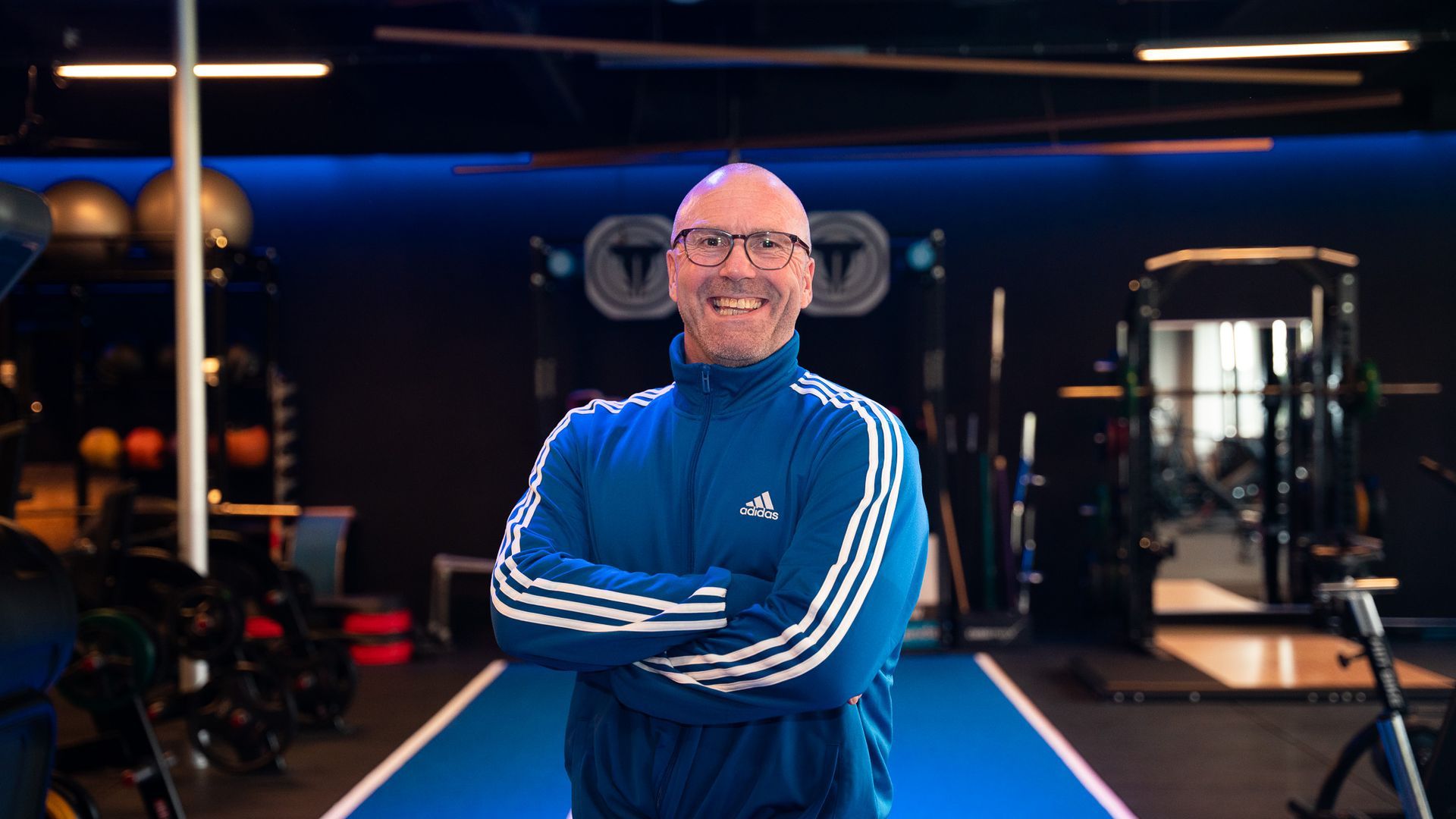 Man in blauw Adidas-trainingspak lacht in de sportschool, armen over elkaar, gewichten op de achtergrond.