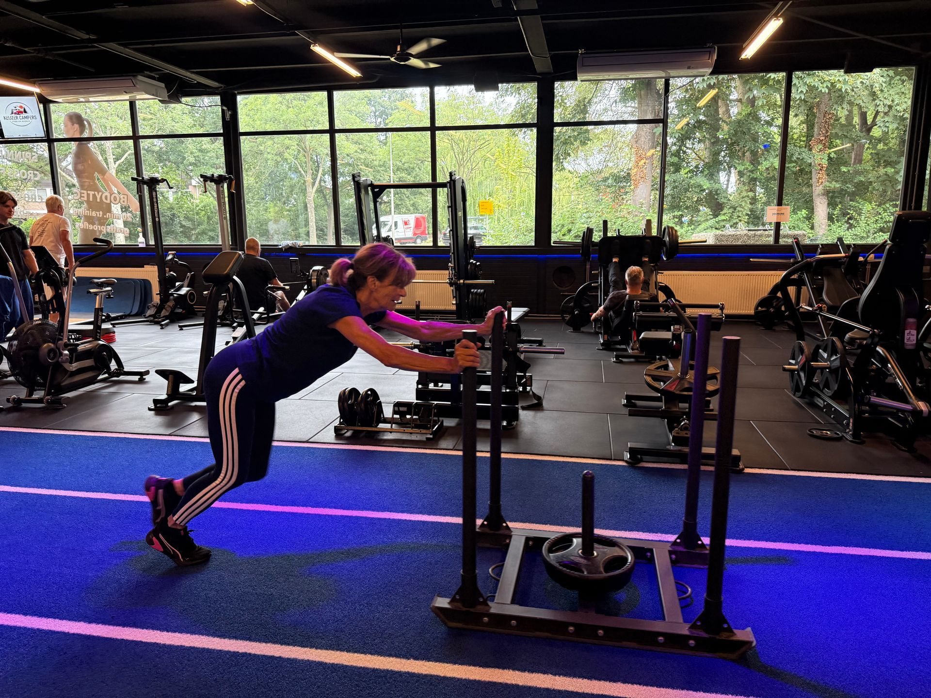 Man op een airbike, lachend met een hometrainer, in de sportschool. Blauw en zwart domineren.
