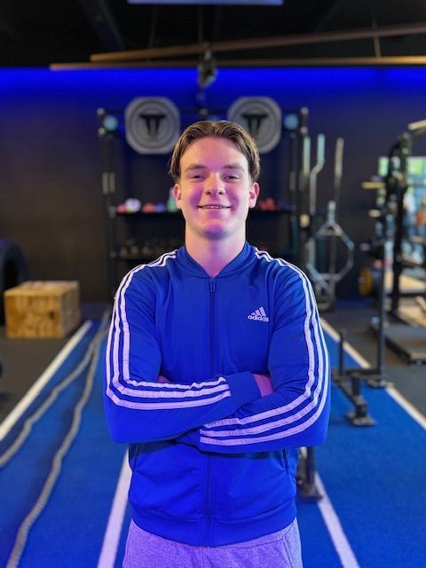Man in een blauwe Adidas-jas met de armen over elkaar in een sportschool.