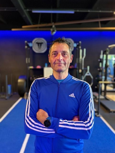Man in een blauw Adidas trainingsjack, met de armen over elkaar, in een sportschool.