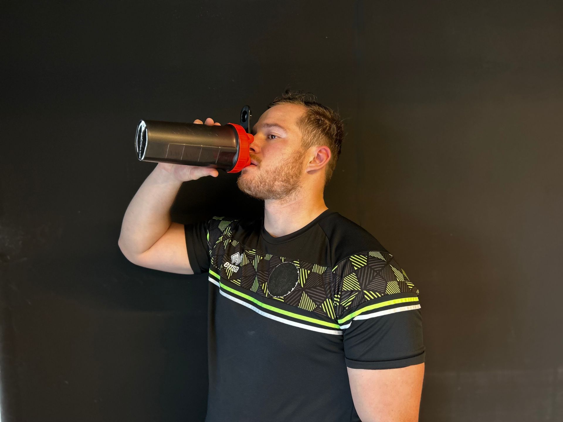 Man drinkt uit een shakerfles, zwarte achtergrond. Hij draagt een zwart-groen sportshirt.