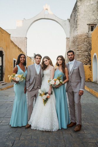 Wedding party poses in courtyard; bride in white gown, bridesmaids in blue, groomsmen in gray suits, holding bouquets.
