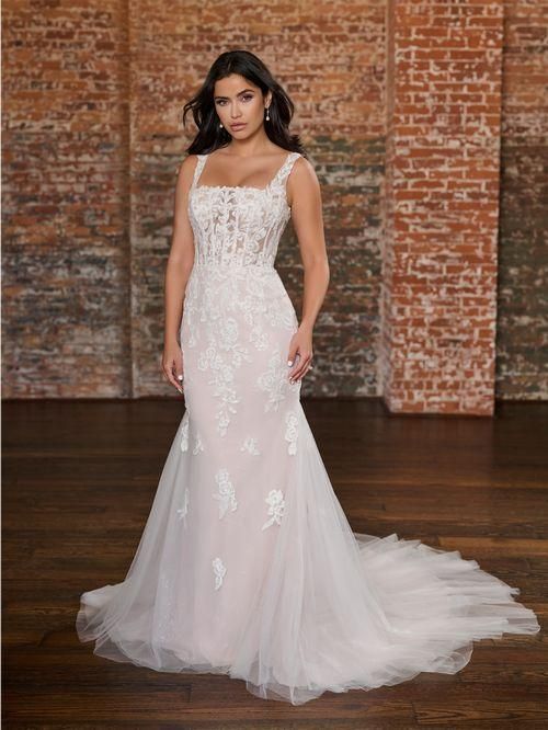 Woman in a white wedding dress with long sleeves, a slit, and a train, standing against a brick wall.