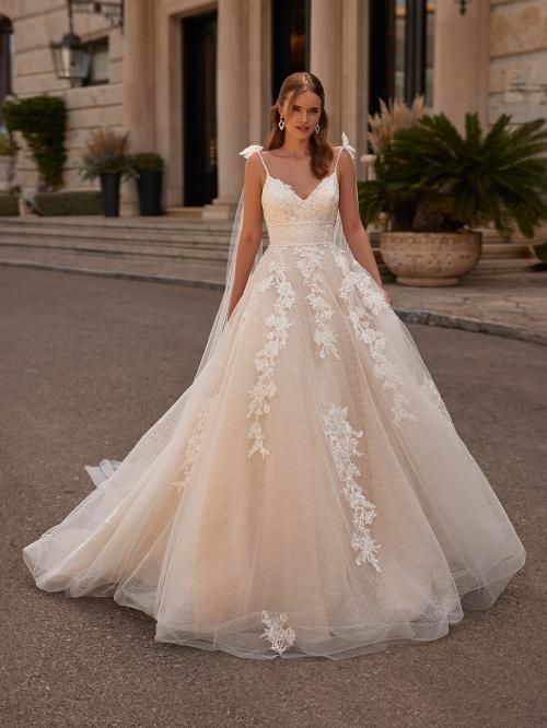 Woman in a light beige wedding dress with lace details and a long train, standing outdoors.