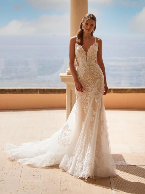 Woman in white lace wedding dress with train, outdoors, smiling, leaning against pillar.