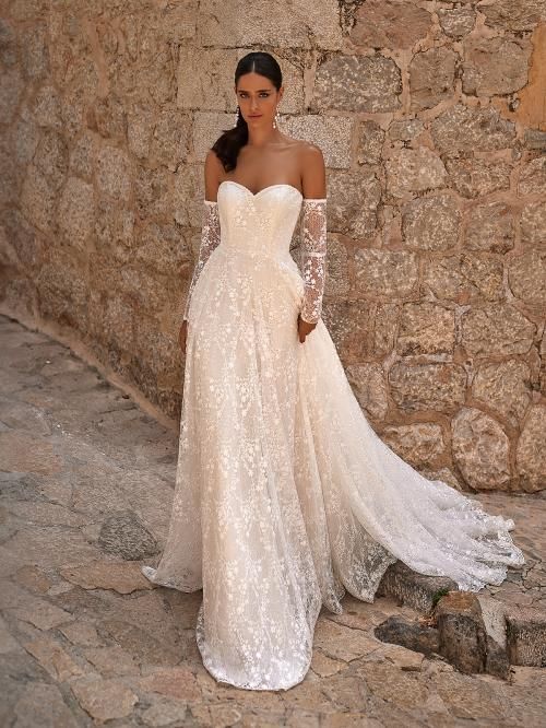 Woman in a white strapless wedding dress with a long train, standing against a stone wall.
