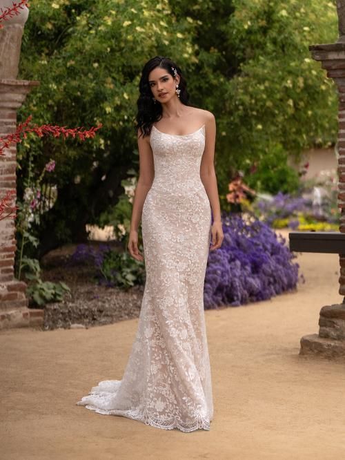 Woman in a white, beaded, one-shoulder wedding dress, standing outdoors in a garden.