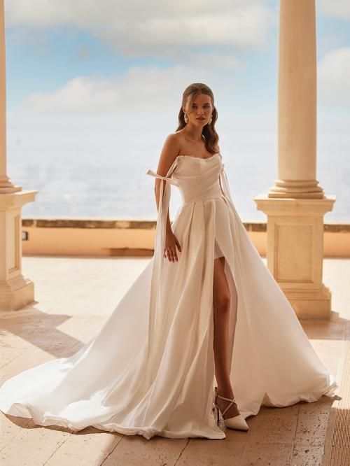 Woman in a white wedding dress with a thigh-high slit, posing outdoors near columns and ocean view.