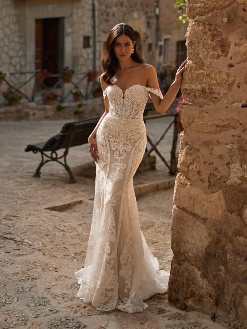 Woman in a lace wedding dress, leaning against a stone wall in a courtyard.