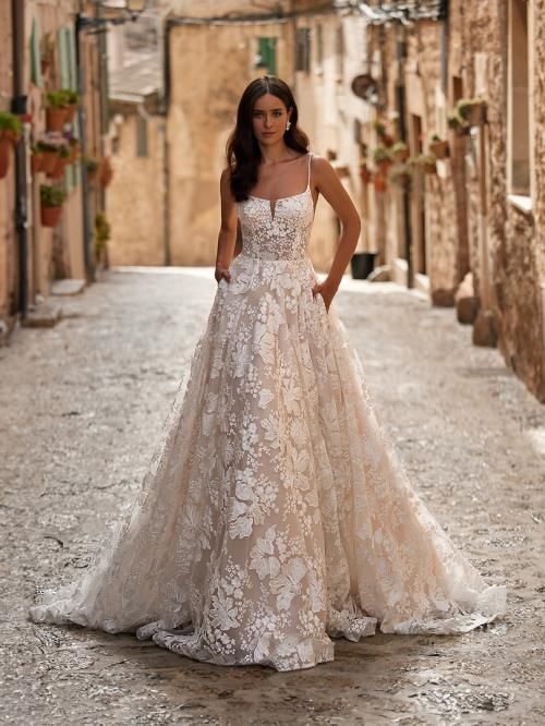 Woman in a wedding dress with floral lace, standing on a cobblestone street, hands in pockets.