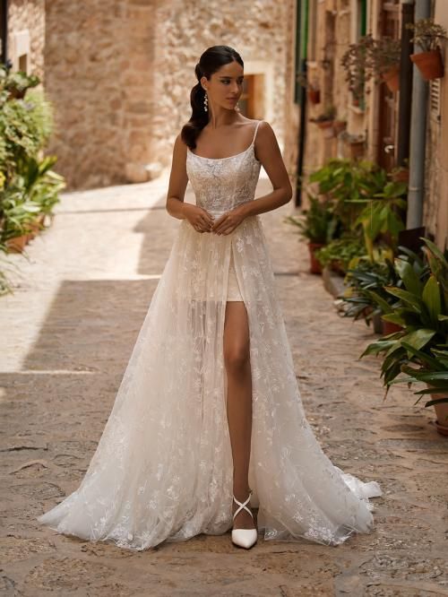 Woman in a wedding dress with a thigh-high slit, standing in a narrow stone alleyway lined with plants.