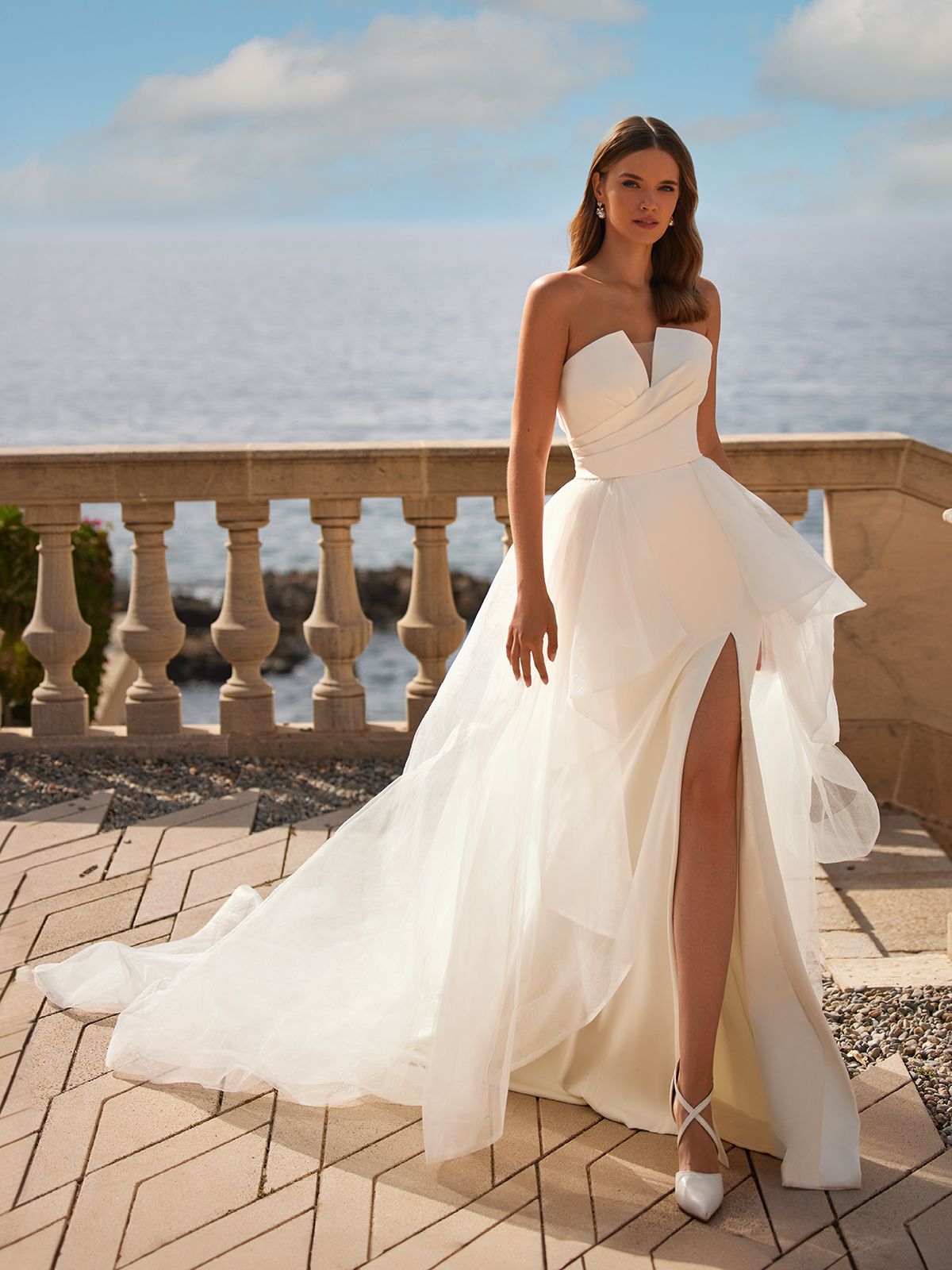 Woman in a strapless white wedding dress with a high slit, standing on a balcony overlooking the ocean.