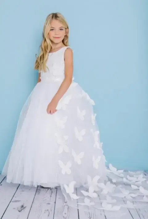 Blond girl in white dress with butterfly accents, posing against a light blue wall.