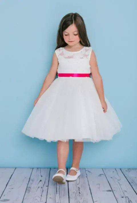 Girl in white dress with pink sash, standing on a wooden floor against a blue background.