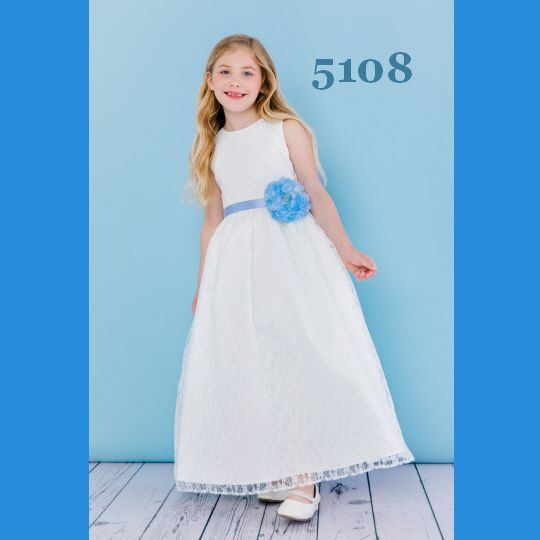 Girl in white dress with blue flower and belt smiles in front of a blue wall.