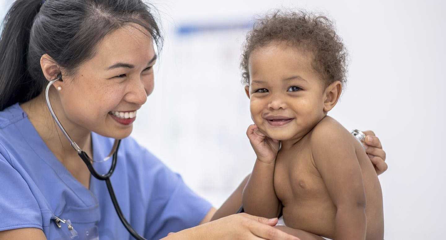 Doctor in blue coat smiling and examining a smiling child with a stethoscope in a clinic