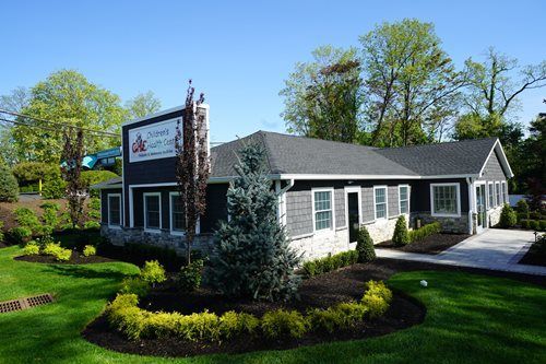 Landscaped gray office building with white trim and sign, surrounded by green trees and lawn