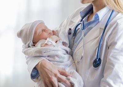 Doctor holding a sleeping newborn wrapped in a white blanket and cap, with a stethoscope around the neck