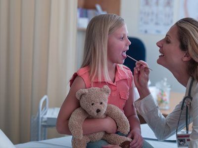 Doctor examining a young girl’s throat while she holds a teddy bear in a clinic