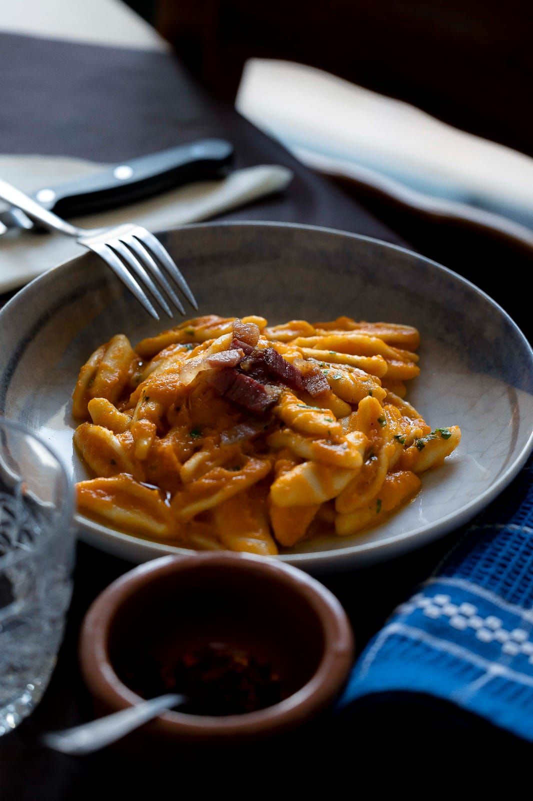 Un piatto di pasta al sugo di pomodoro e una forchetta su un tavolo scuro, con una tovaglia a quadri blu nelle vicinanze.