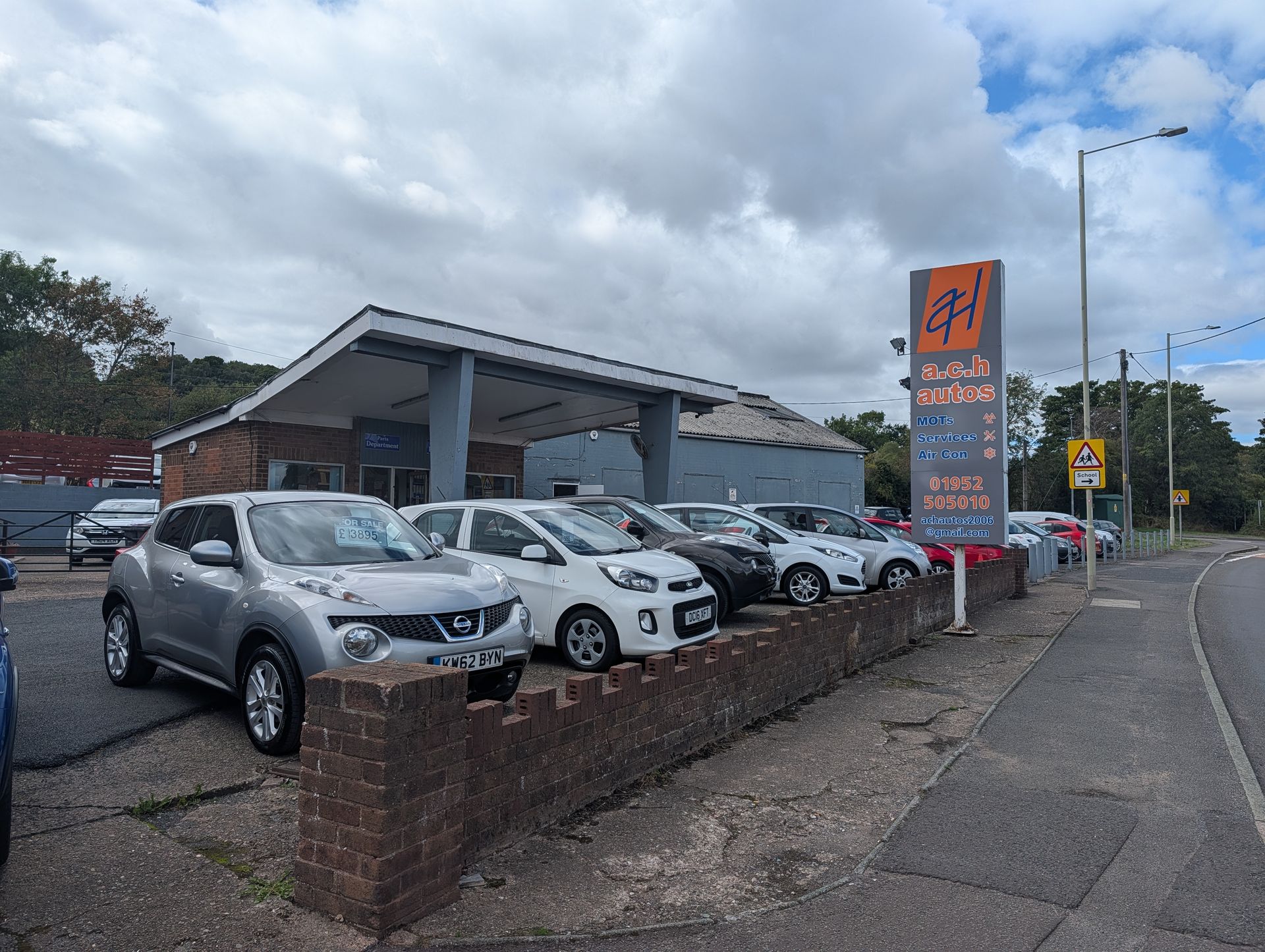 Car lot with several cars, a gray SUV in front, and an orange sign beside a roadside under cloudy skies