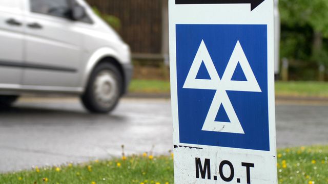 Blue M.O.T. road sign with white symbol beside a passing silver car on a roadside
