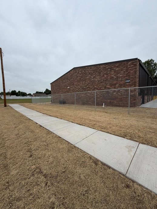 Concrete walkway beside a brick building under a cloudy sky, with gravel and open grass around it