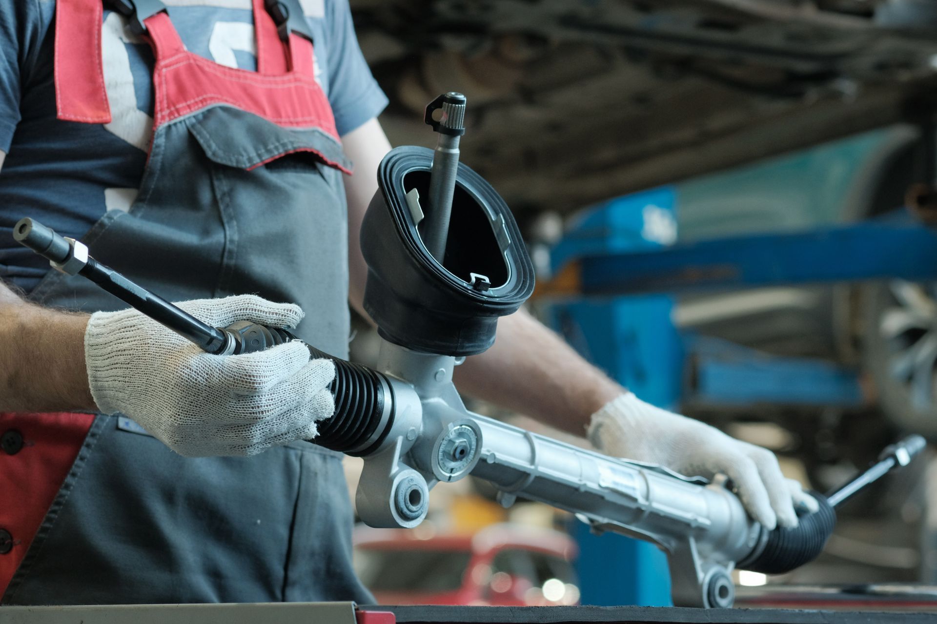 Mechanic holding a car steering rack with gloves on, in a garage setting.